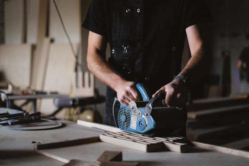 Three professionals in workshop attire reviewing a belt sander at a timber workbench, holding sanding belts and calipers